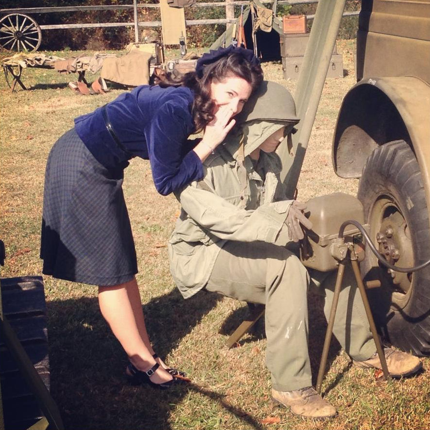 Woman in a blue sweater and skirt leaning over a man in military uniform next to a vehicle.